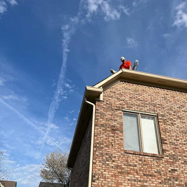 A man in an orange shirt sits on the edge of a brick house roof under a bright blue sky with wispy clouds, possibly during a roof replacement or repair.