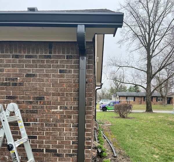 Close-up of a newly installed dark brown gutter and downspout on a brick house, part of a gutter guard installation project. A ladder leans against the brick wall. Green lawn and trees in the background.
