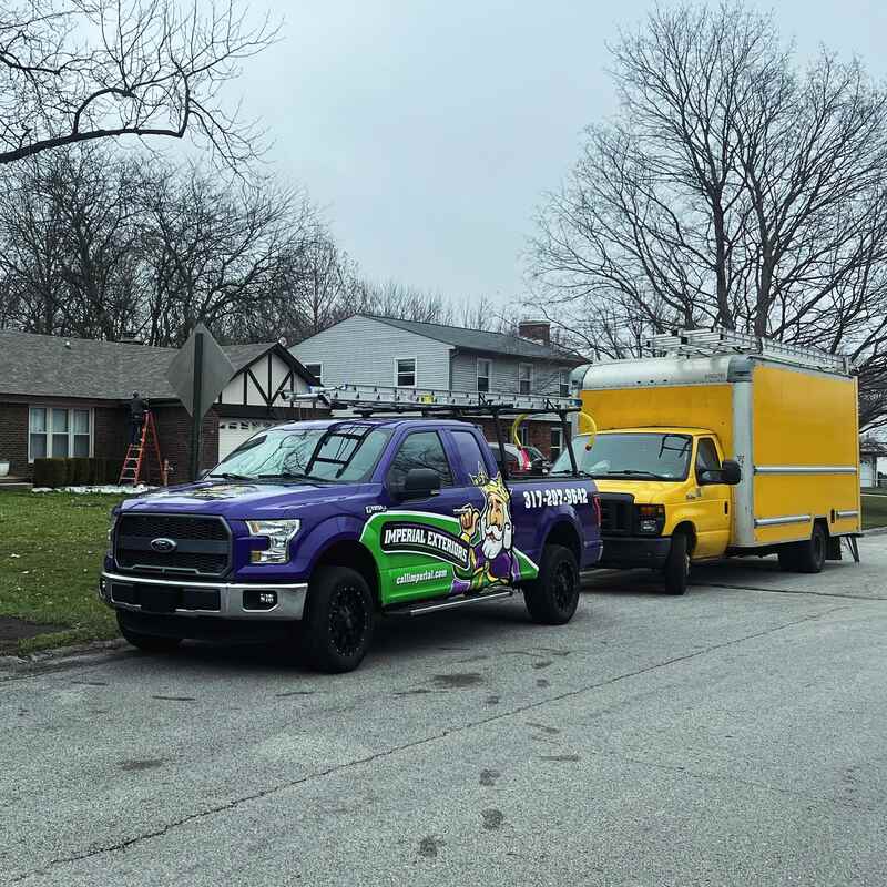 Imperial Exteriors truck and trailer parked on a residential street, ready for roof replacement after storm damage. The truck is purple and green, with the company's phone number: 317-207-3642.