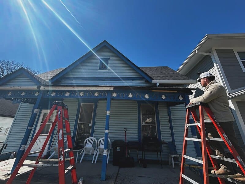 Man on ladder inspecting roof damage on a light blue Victorian house. Roof replacement work is underway after storm damage.